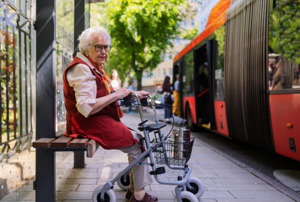 Senior woman in city, sitting on a bus stop, waiting for public transport, rollator next to her.