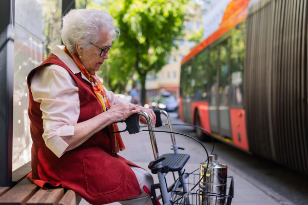 Senior woman in city, sitting on a bus stop, waiting for public transport, rollator next to her.