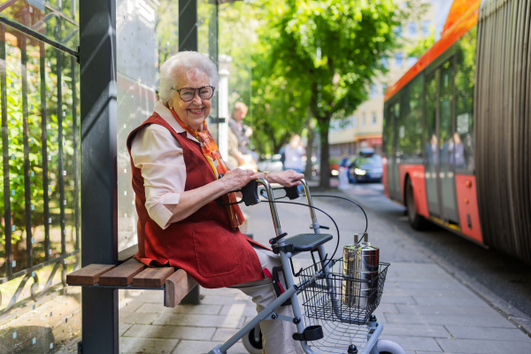 Senior woman in city, sitting on a bus stop, waiting for public transport, rollator next to her.