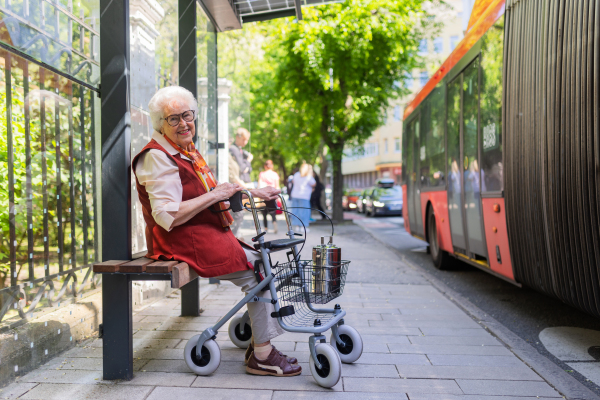 Senior woman in city, sitting on a bus stop, waiting for public transport, rollator next to her.