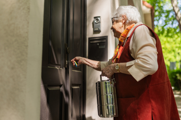 Elderly woman in front of her house, unlocking the gate with key. Holding lunch container with a meal from canteen restaurant.