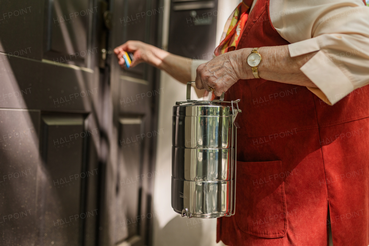 Elderly woman in front of her house, unlocking the gate with key. Holding lunch container with a meal from canteen restaurant.