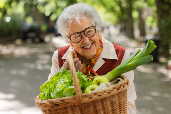 Senior woman going home from the supermarket, walking in the city, holding basket full of groceries.