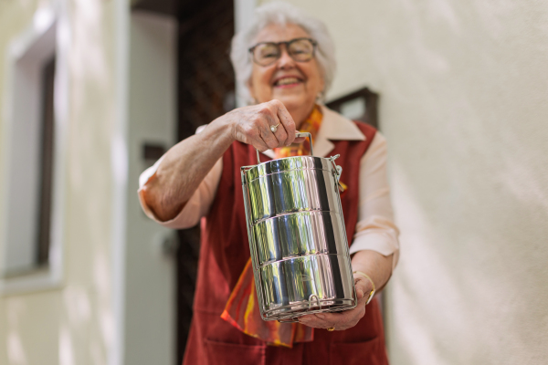 Smiling elderly woman in front of her house, holding lunch container with a meal from canteen or restaurant.