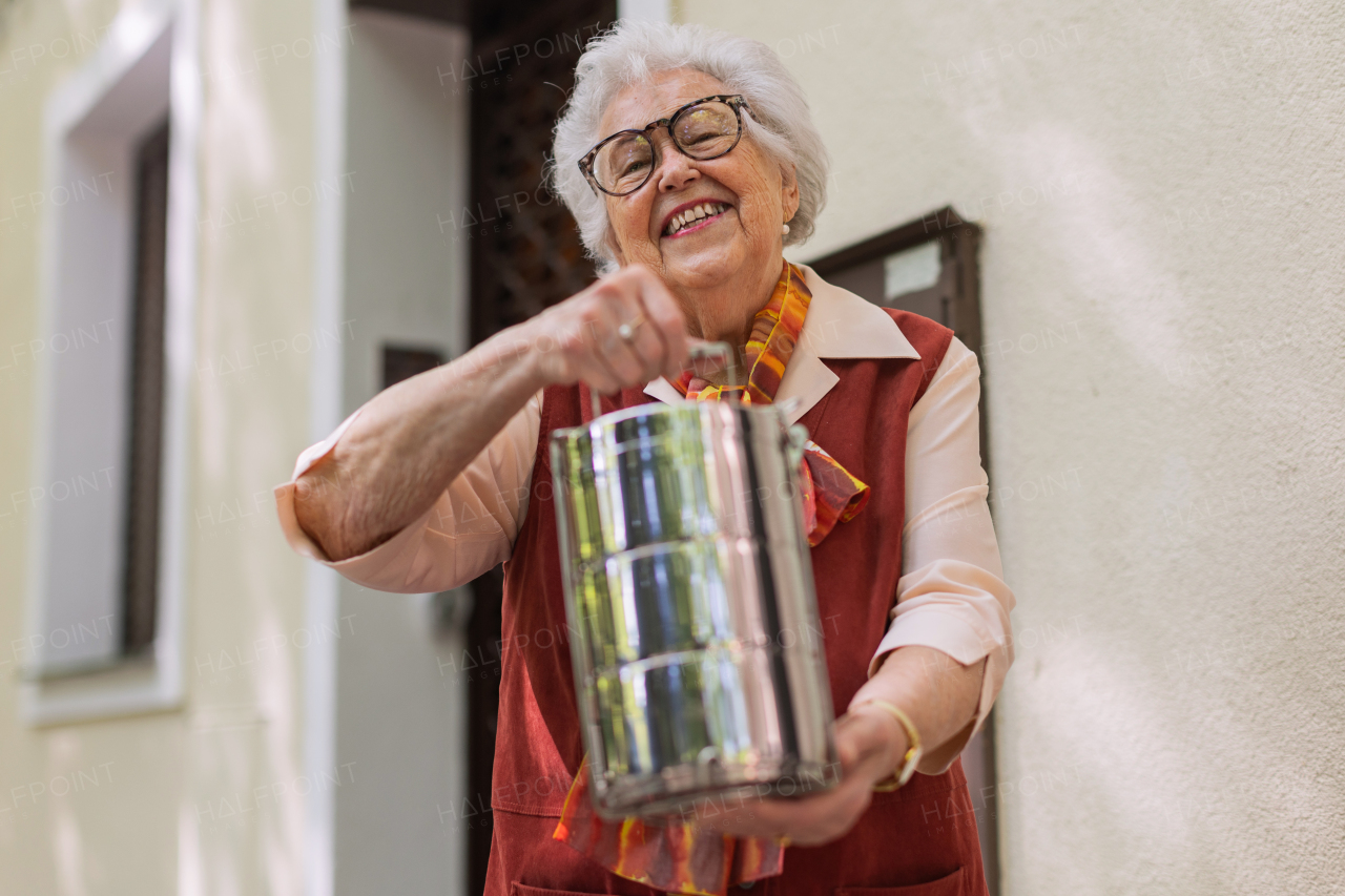 Smiling elderly woman in front of her house, holding lunch container with a meal from canteen or restaurant.