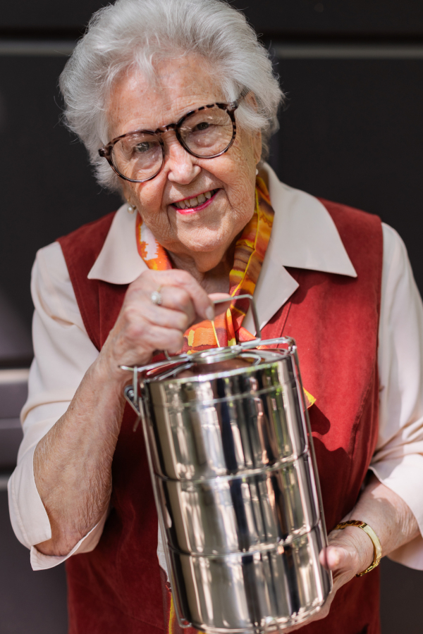 Smiling elderly woman in front of her house, holding lunch container with a meal from canteen or restaurant.