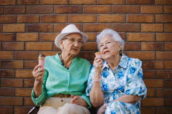 Portrait of two senior female friends in the city, eating ice cream on a hot summer day. Elderly ladies on summer vacation in the city. Pensioners on group trip. Concept of senior vacation and travel groups, clubs.