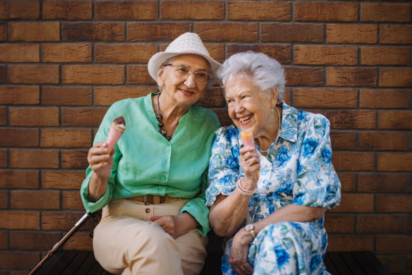 Portrait of two senior female friends in the city, eating ice cream on a hot summer day. Elderly ladies on summer vacation in the city. Pensioners on group trip. Concept of senior vacation and travel groups, clubs.