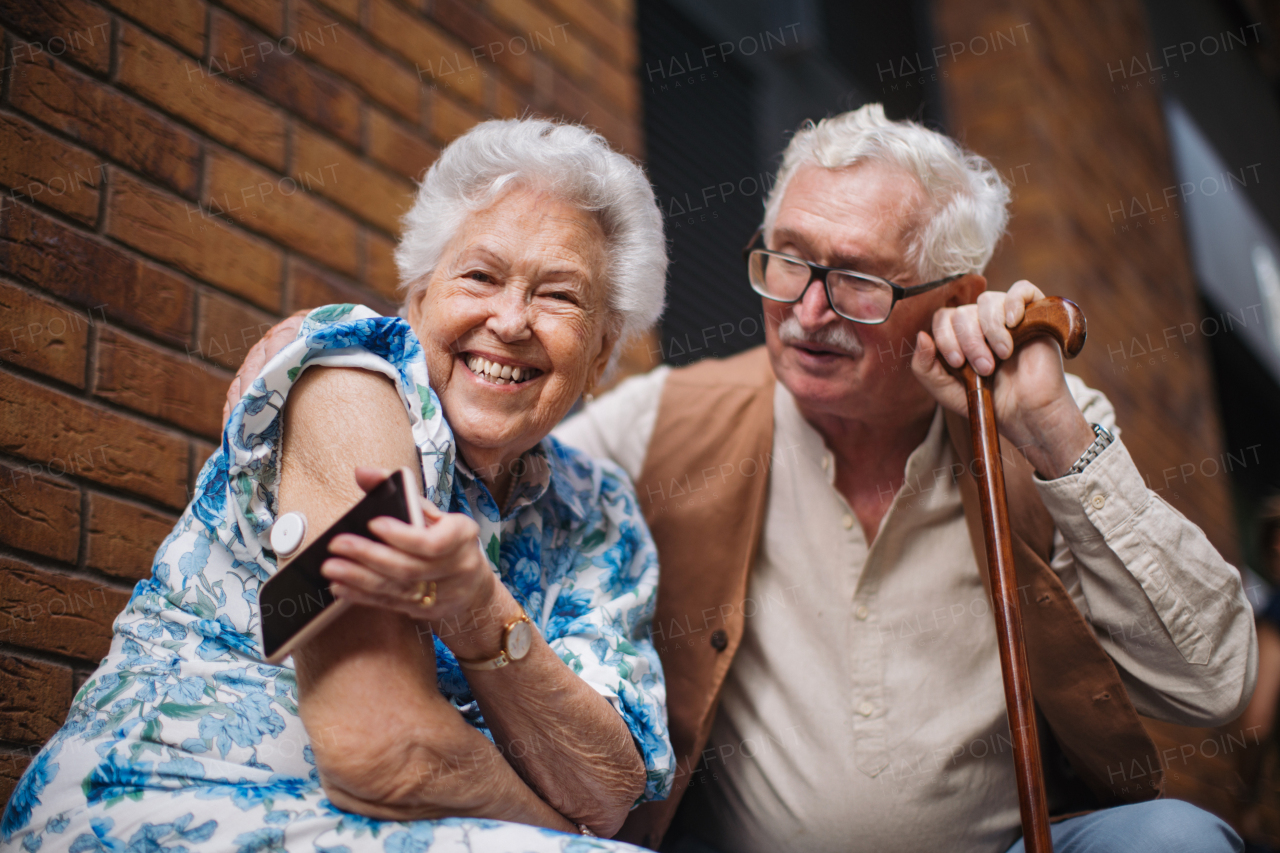 Chronic illnes in elderly couple. Diabetic senior woman checking blood glucose level with continuous glucose monitor outdoors in the city. Elderly husband waiting while his chronically ill wife connecting her CGM with smarphone.