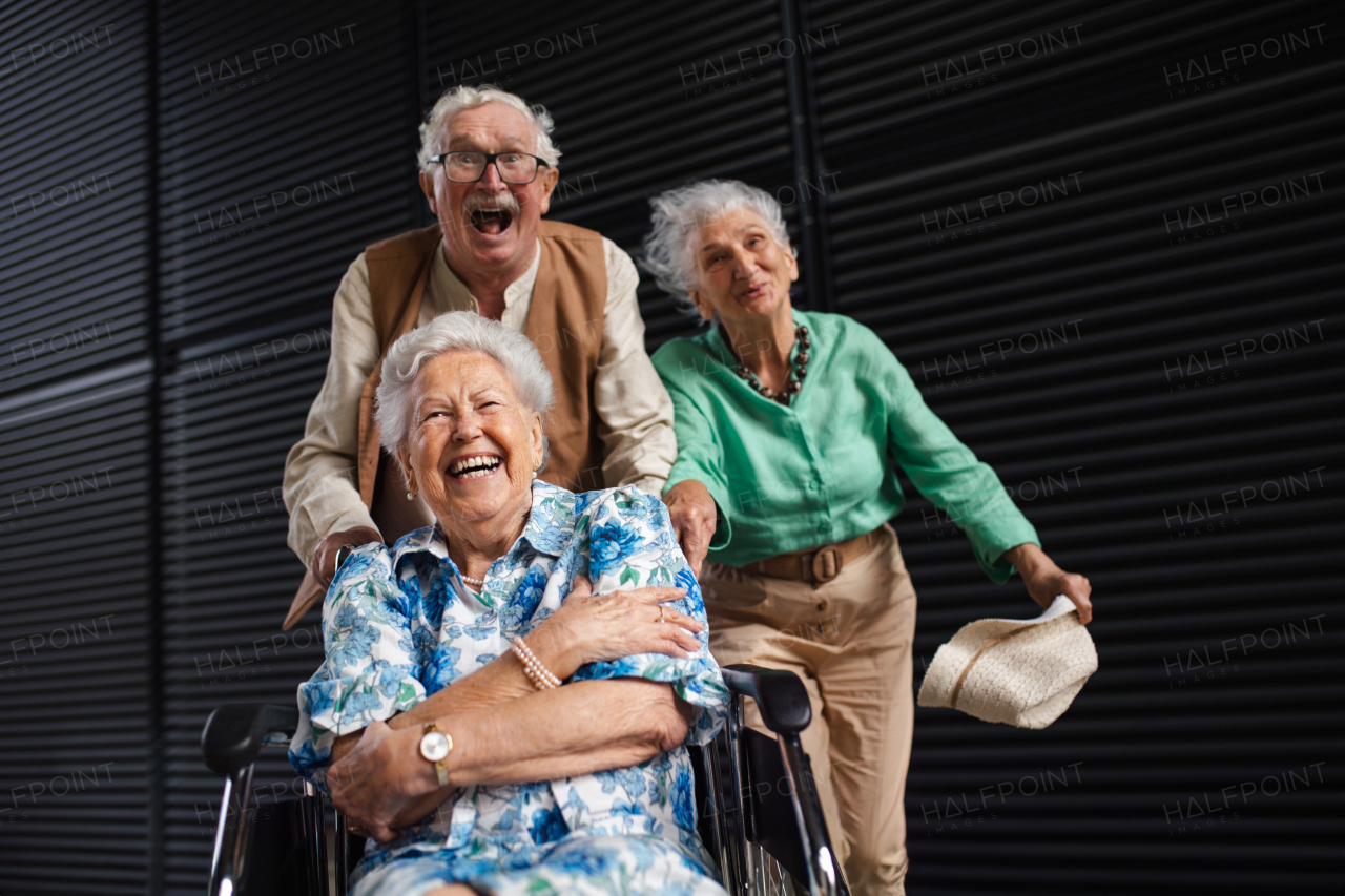 Portrait of three senior friends in the city, having fun. Funny pensioners pushing one of friend on wheelchair very fast. Concept of active retirement. Portrait with copy space on black backgroun.