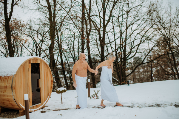 Senior woman in towel with her husband coming out from outdoor sauna during a cold winter day.