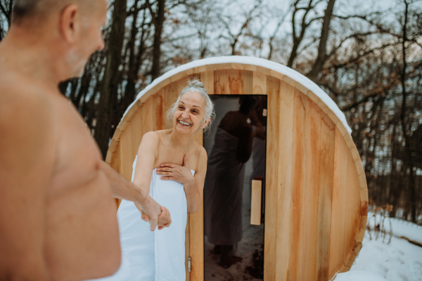 Senior woman in towel with her husband coming out from outdoor sauna during a cold winter day.