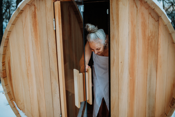 Senior woman in towel coming out from outdoor sauna during a cold winter day.