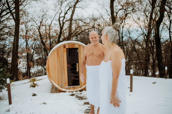 Senior woman in towel with her husband coming out from outdoor sauna during a cold winter day.