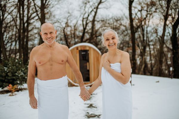 Senior couple in love standing in towels in front of outdoor wooden sauna during cold winter day.