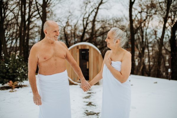 Senior couple in love standing in towels in front of outdoor wooden sauna during cold winter day.