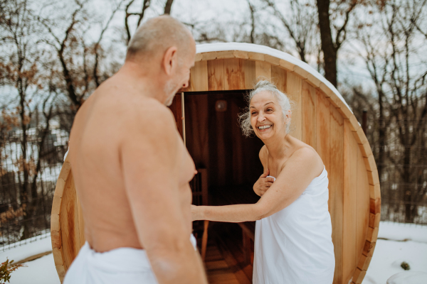 Senior woman in towel with her husband coming out from outdoor sauna during a cold winter day.