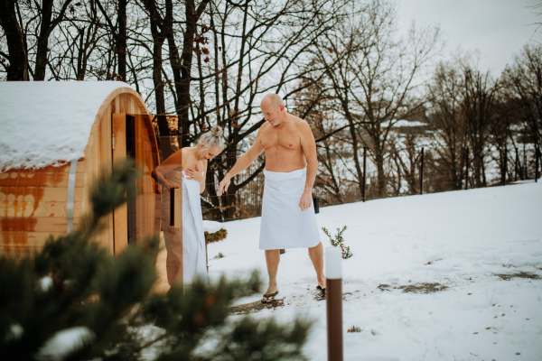 Senior woman in towel with her husband coming out from outdoor sauna during a cold winter day.