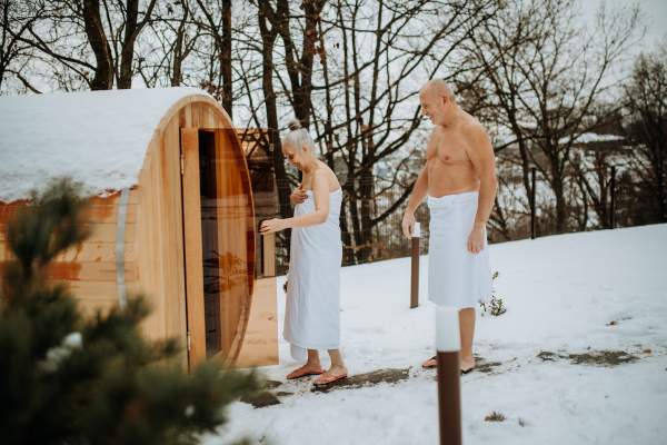 Senior woman in towel with her husband coming out from outdoor sauna during a cold winter day.