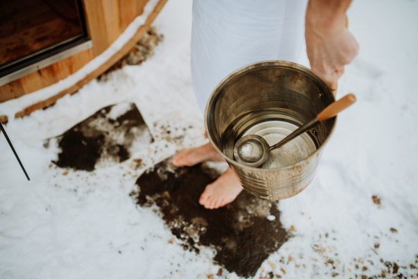 Close-up of senior man holding bucket of water and entering in outdoor wooden sauna during cold winter day.