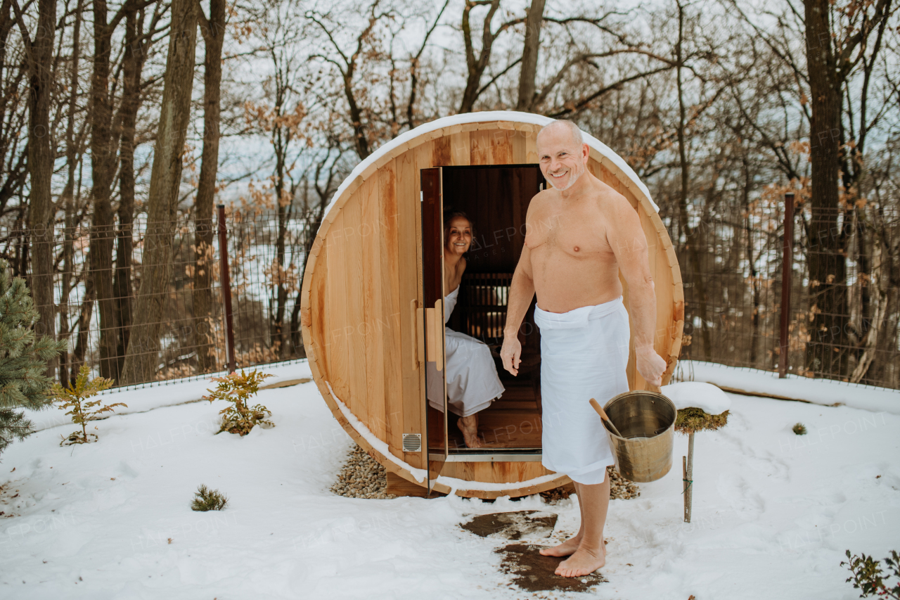 Senior woman in towel with her husband coming out from outdoor sauna during a cold winter day.