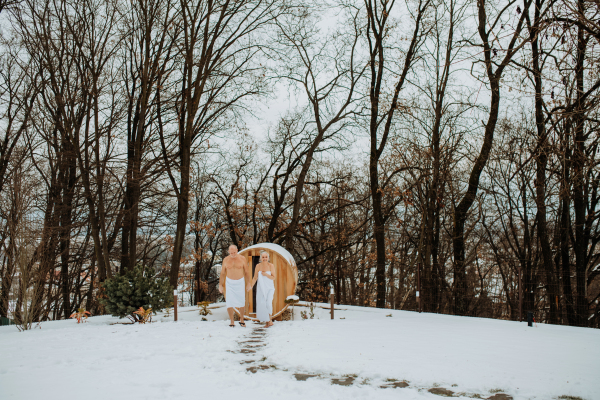 Senior woman in towel with her husband coming out from outdoor sauna during a cold winter day.