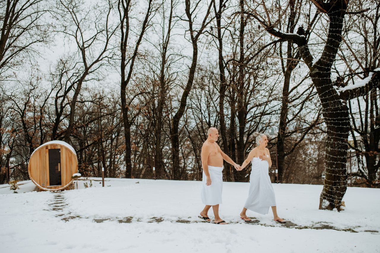 Senior woman in towel with her husband coming out from outdoor sauna during a cold winter day.