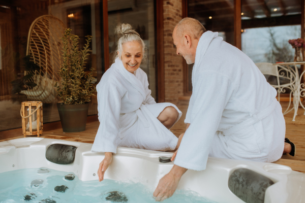 Senior couple in a bathrobe checking temperature in outdoor hot tub, preparing for bathing with his wife.