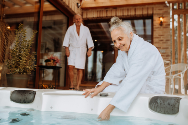 Senior woman in a bathrobe checking temperature in outdoor hot tub, preparing for bathing with her husband.