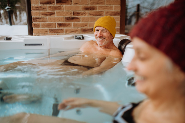 Senior couple in knitted cap enjoying together outdoor bathtub at their terrace during a cold winter day.