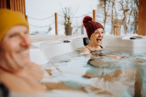 Senior couple in knitted cap enjoying together outdoor bathtub at their terrace during a cold winter day.