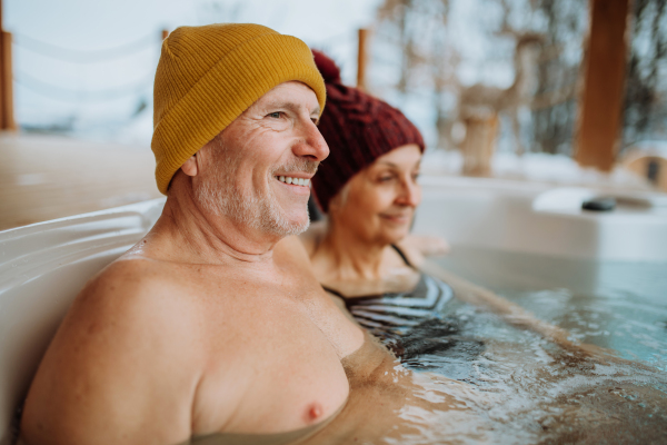 Senior couple in knitted cap enjoying together outdoor bathtub at their terrace during a cold winter day.