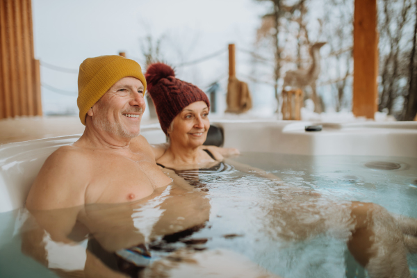 Senior couple in knitted cap enjoying together outdoor bathtub at their terrace during a cold winter day.