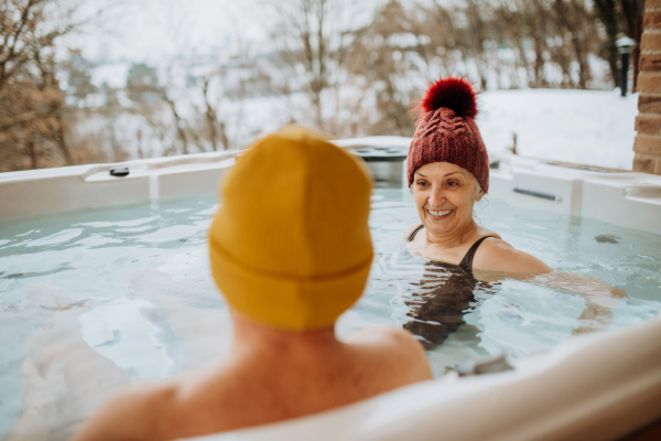 Senior couple in knitted cap enjoying together outdoor bathtub at their terrace during a cold winter day.