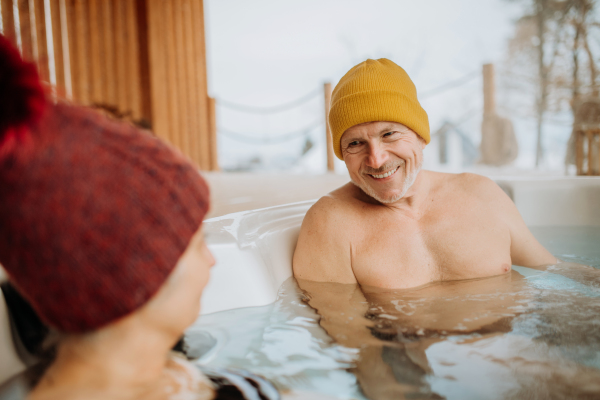 Senior couple in knitted cap enjoying together outdoor bathtub at their terrace during a cold winter day.