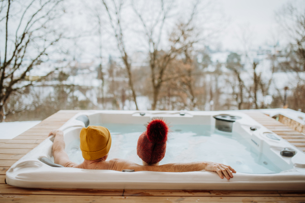 Senior couple in knitted cap enjoying together outdoor bathtub at their terrace during a cold winter day.