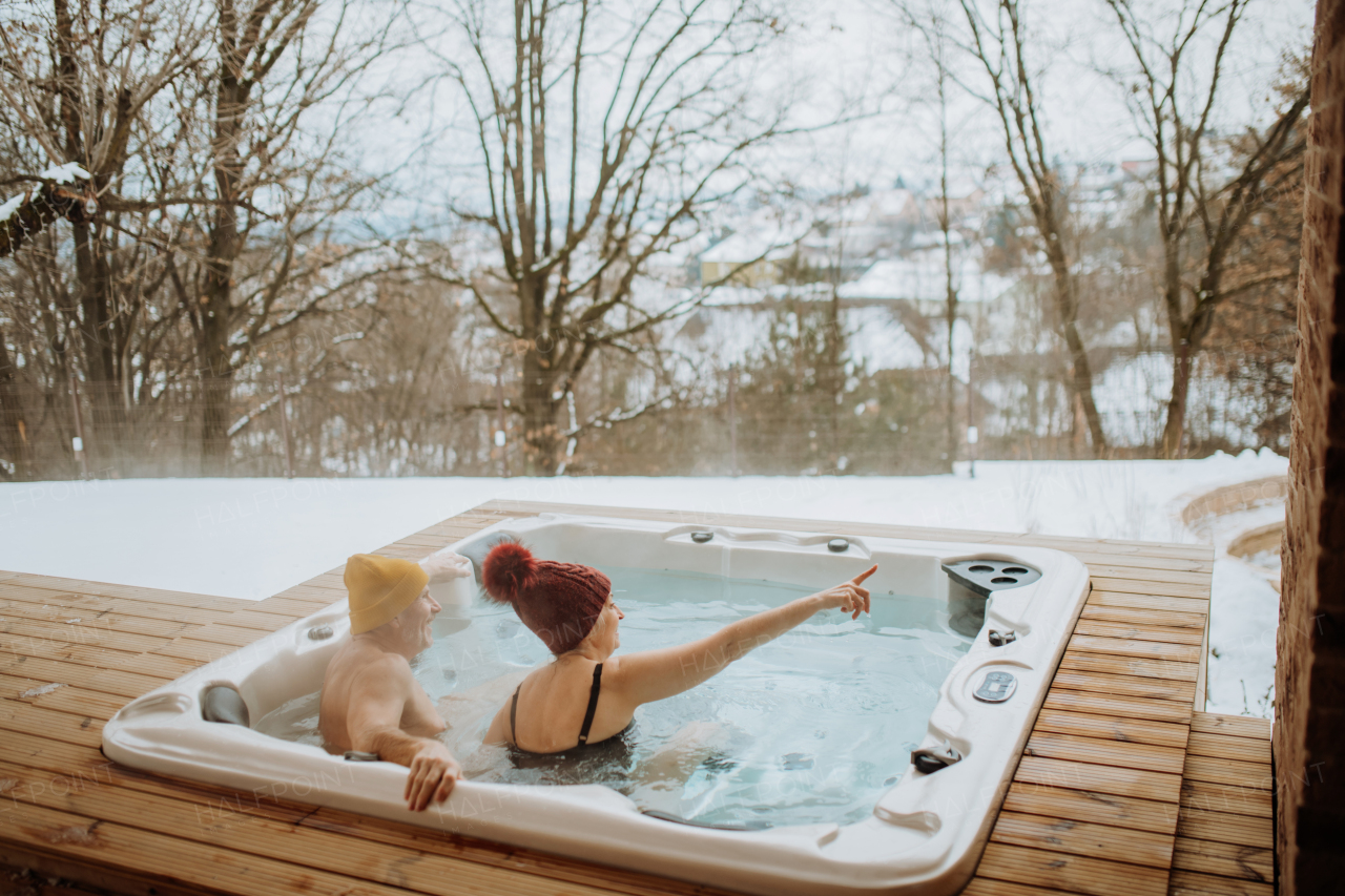 Senior couple in knitted cap enjoying together outdoor bathtub and view at their terrace during a cold winter day.