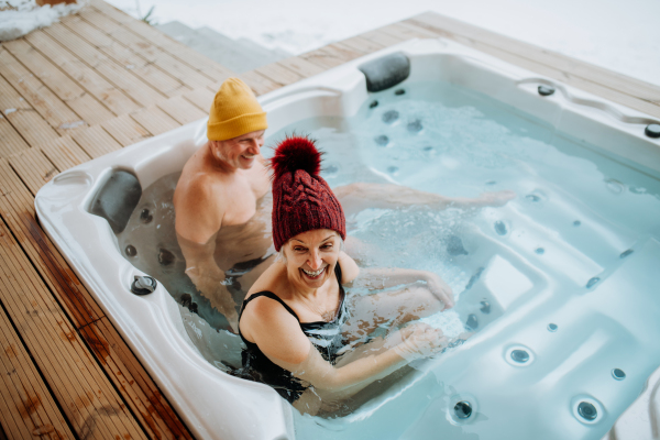 Senior couple in knitted cap enjoying together outdoor bathtub at their terrace during a cold winter day.