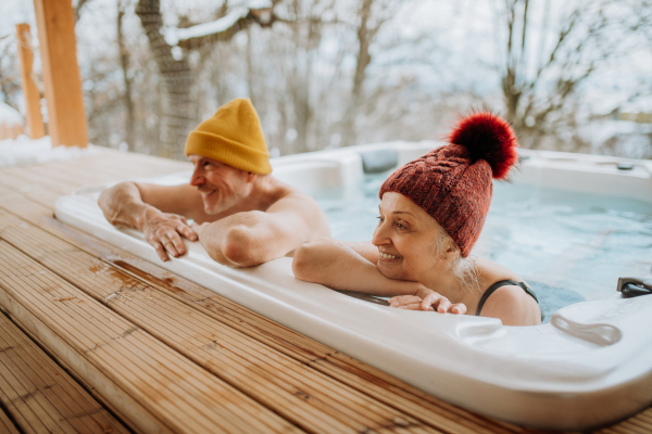 Senior couple in knitted cap enjoying together outdoor bathtub at their terrace during a cold winter day.