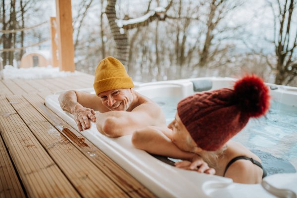 Senior couple in knitted cap enjoying together outdoor bathtub at their terrace during a cold winter day.