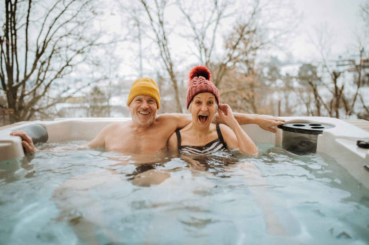 Senior couple in knitted cap enjoying together outdoor bathtub at their terrace during a cold winter day.