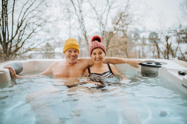 Senior couple in knitted cap enjoying together outdoor bathtub at their terrace during a cold winter day.