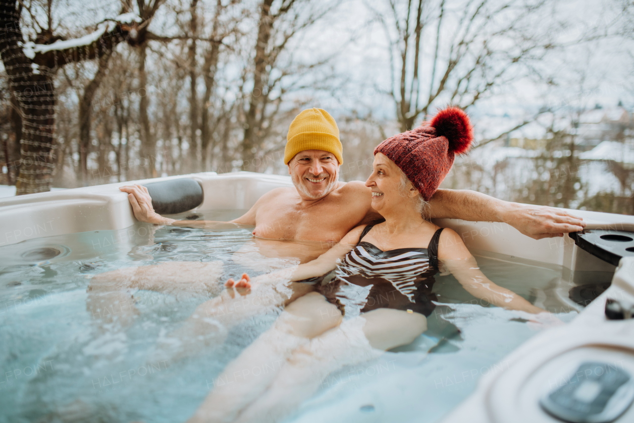 Senior couple in knitted cap enjoying together outdoor bathtub at their terrace during a cold winter day.