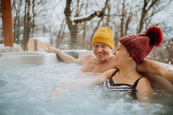 Senior couple in knitted cap enjoying together outdoor bathtub at their terrace during a cold winter day.