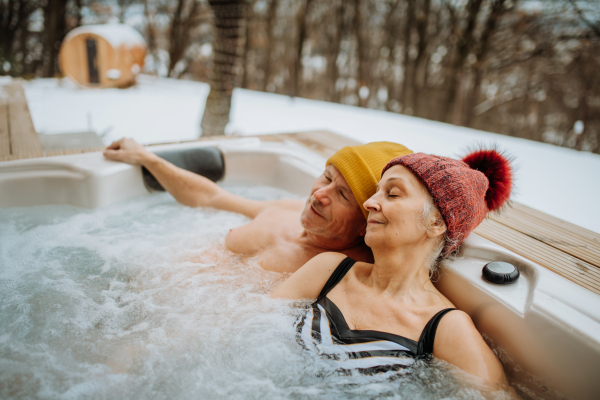 Senior couple in knitted cap enjoying together outdoor bathtub at their terrace during a cold winter day.