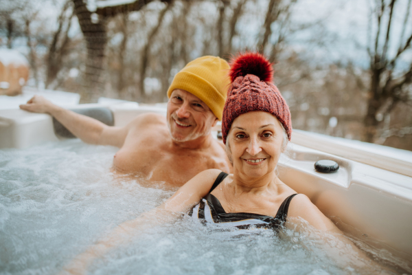 Senior couple in knitted cap enjoying together outdoor bathtub at their terrace during a cold winter day.