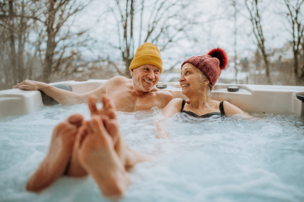 Senior couple in knitted cap enjoying together outdoor bathtub at their terrace during a cold winter day.