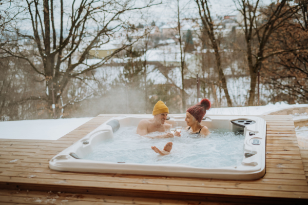 Senior couple in knitted cap enjoying together outdoor bathtub with glass of wine at their terrace during a cold winter day.