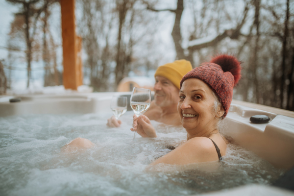 Senior couple in kintted cap enjoying together outdoor bathtub and enjoying glass of wine at their terrace during a cold winter day.