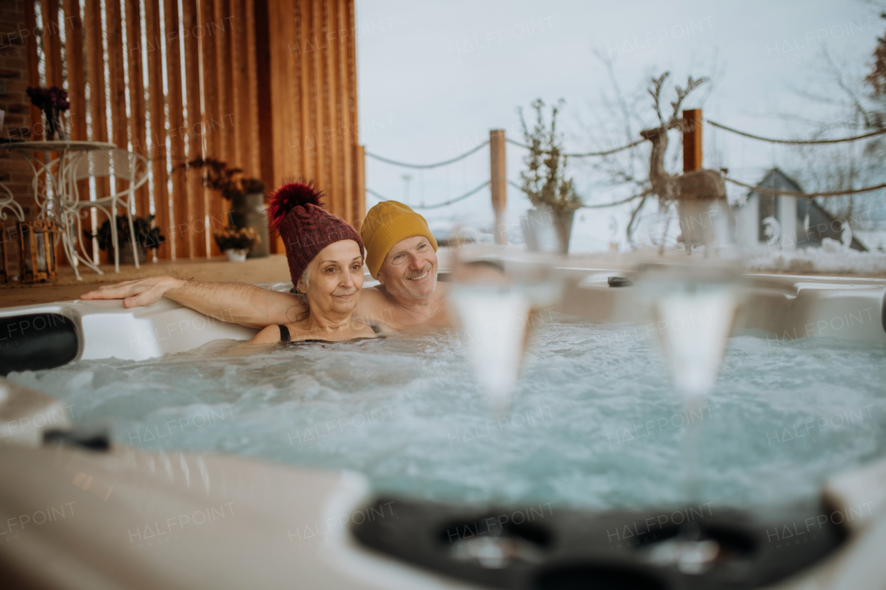 Senior couple in knitted cap enjoying together outdoor bathtub at their terrace during a cold winter day.